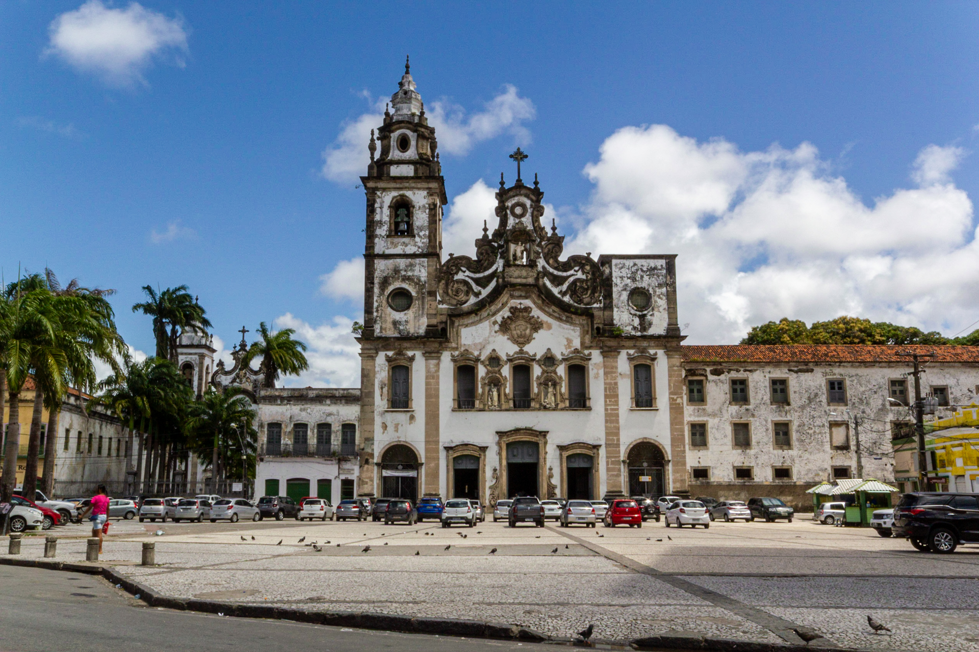 Fachada da Basílica de Nossa Senhora do Carmo de Recife, a sua direita a fachada da edificação do convento, e as esquerda a palmeiras, a sua frente há diversos carros estacionados na calçada.