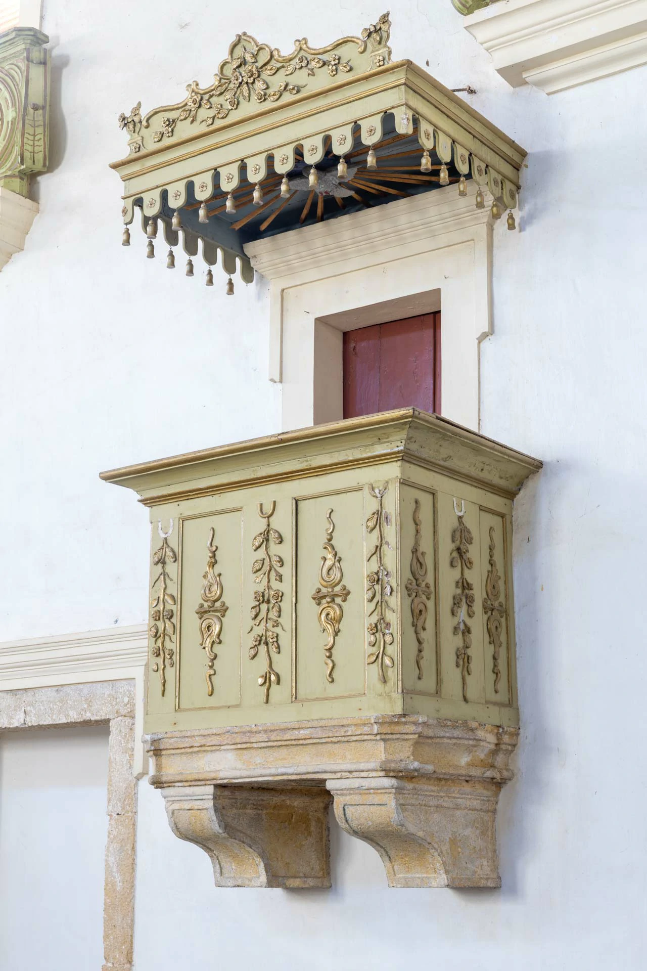 Pulpito da igreja de Nossa Senhora do Monte de madeira com base de pedra natural fixado na parede, formato quadrangular, com adornos barrocos e a imagem do espirito santo no interior do seu topo, a porta é de madeira na cor vermelha.